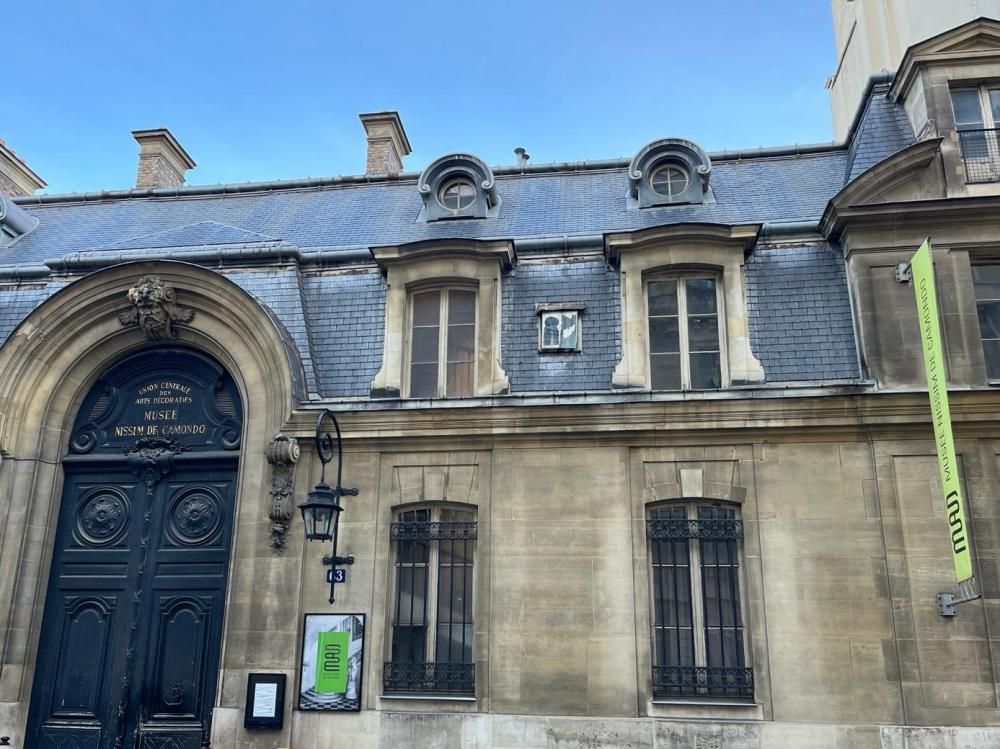 Façade d’un bâtiment historique en pierre, avec une porte bleue cintrée et des fenêtres à barreaux, sous un toit en ardoise.