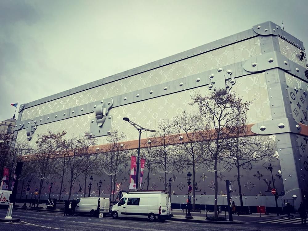 Grand bâtiment moderne gris avec une camionnette blanche garée devant, dans une rue pluvieuse