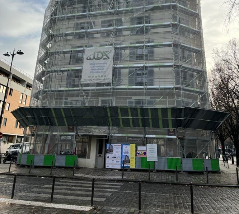 Façade d’un bâtiment en cours de rénovation, avec une bâche verte et des barrières de chantier dans une rue pluvieuse