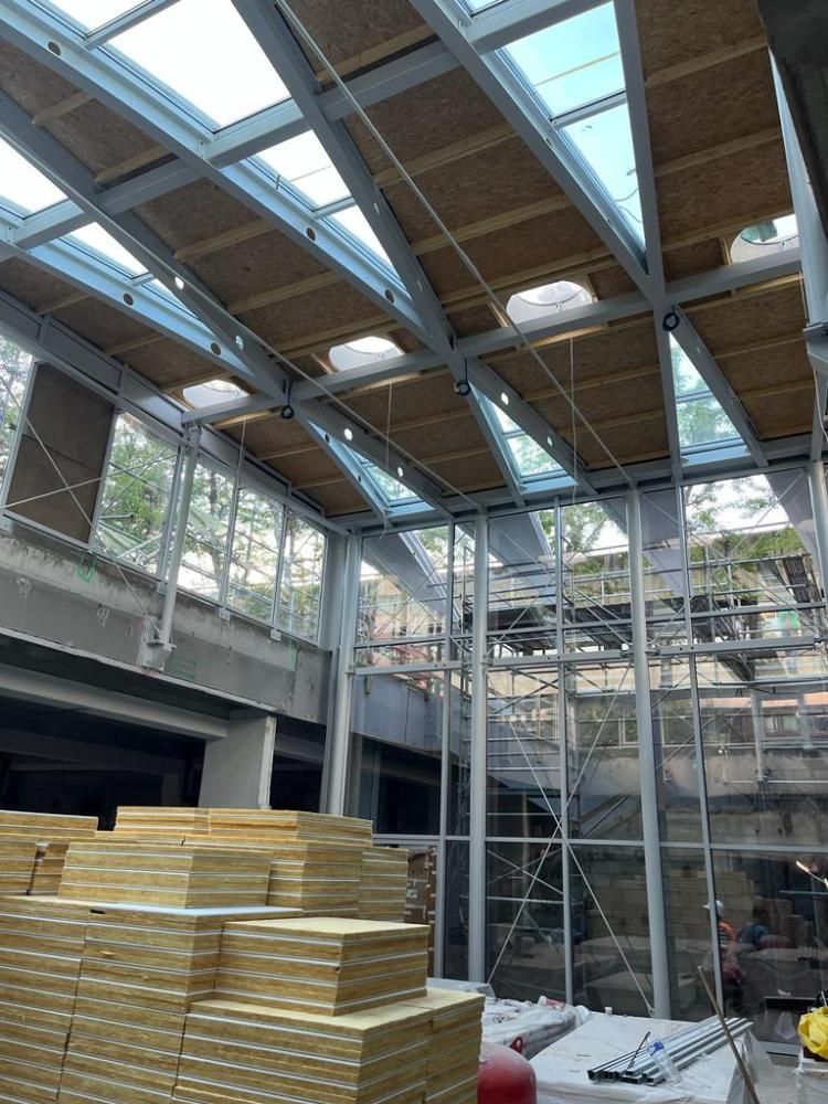 Intérieur d’un chantier avec des poutres en acier apparentes, des puits de lumière et des piles de bois au sol