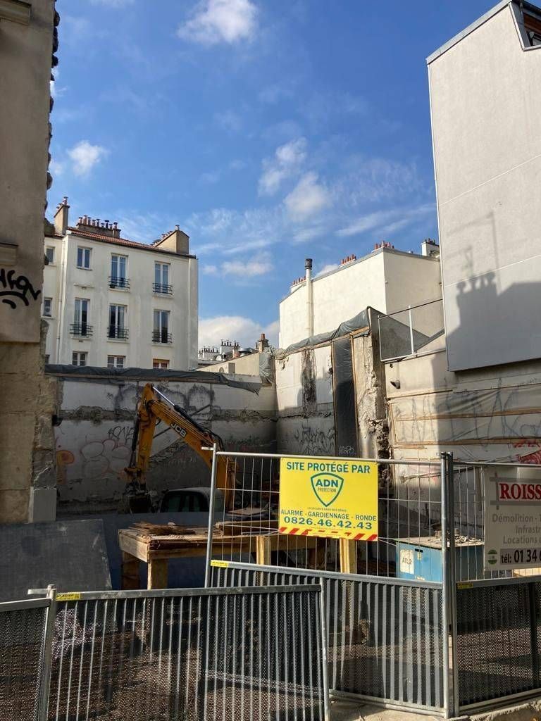 Chantier urbain avec une excavatrice derrière des barrières métalliques, entre des immeubles résidentiels, sous un ciel bleu