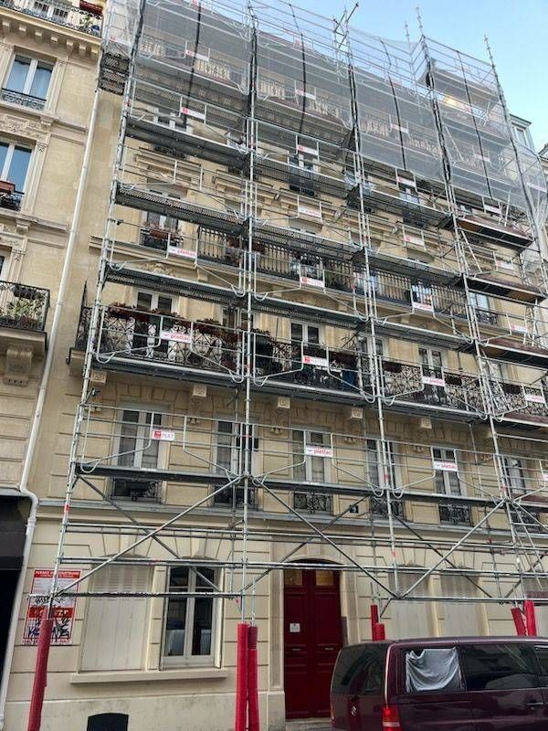 Façade d’un bâtiment en pierre recouverte d’échafaudages, avec une porte rouge et une voiture garée dans une rue de la ville