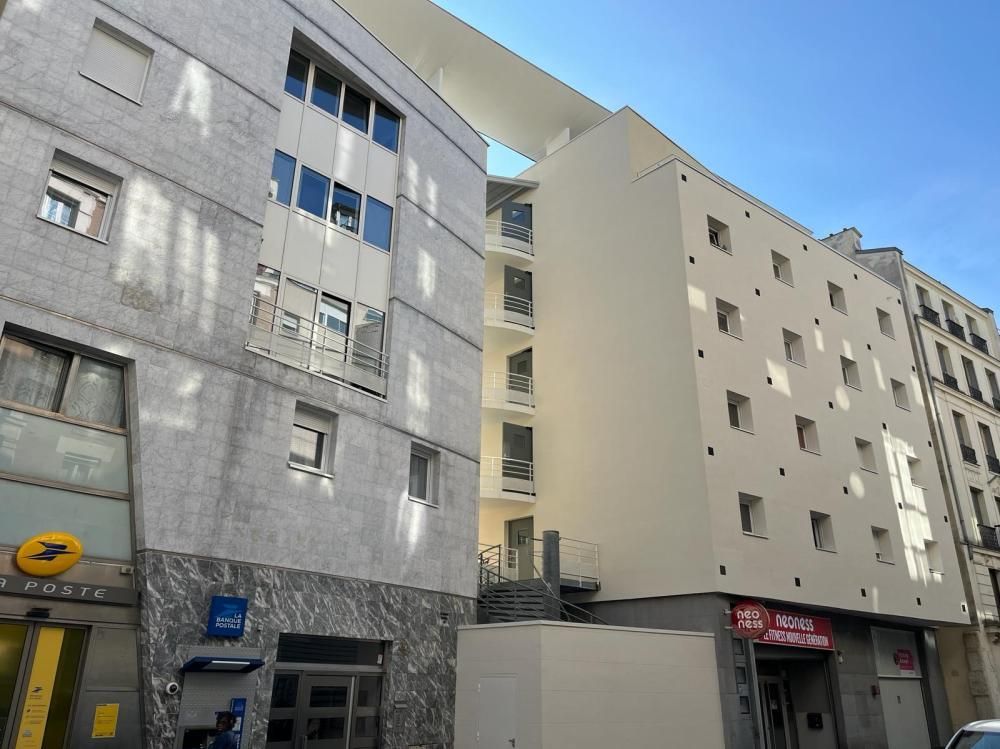 Rue étroite en ville, bordée d’immeubles gris et beiges, sous un ciel bleu