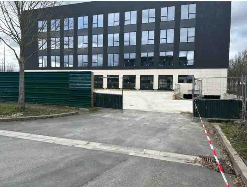 Immeuble de bureaux moderne avec de grandes fenêtres, une allée pavée et des barrières de chantier vertes devant l’entrée