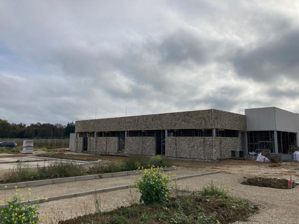 Bâtiment commercial en pierre partiellement construit, sous un ciel nuageux, avec un terrain de gravier à l’avant