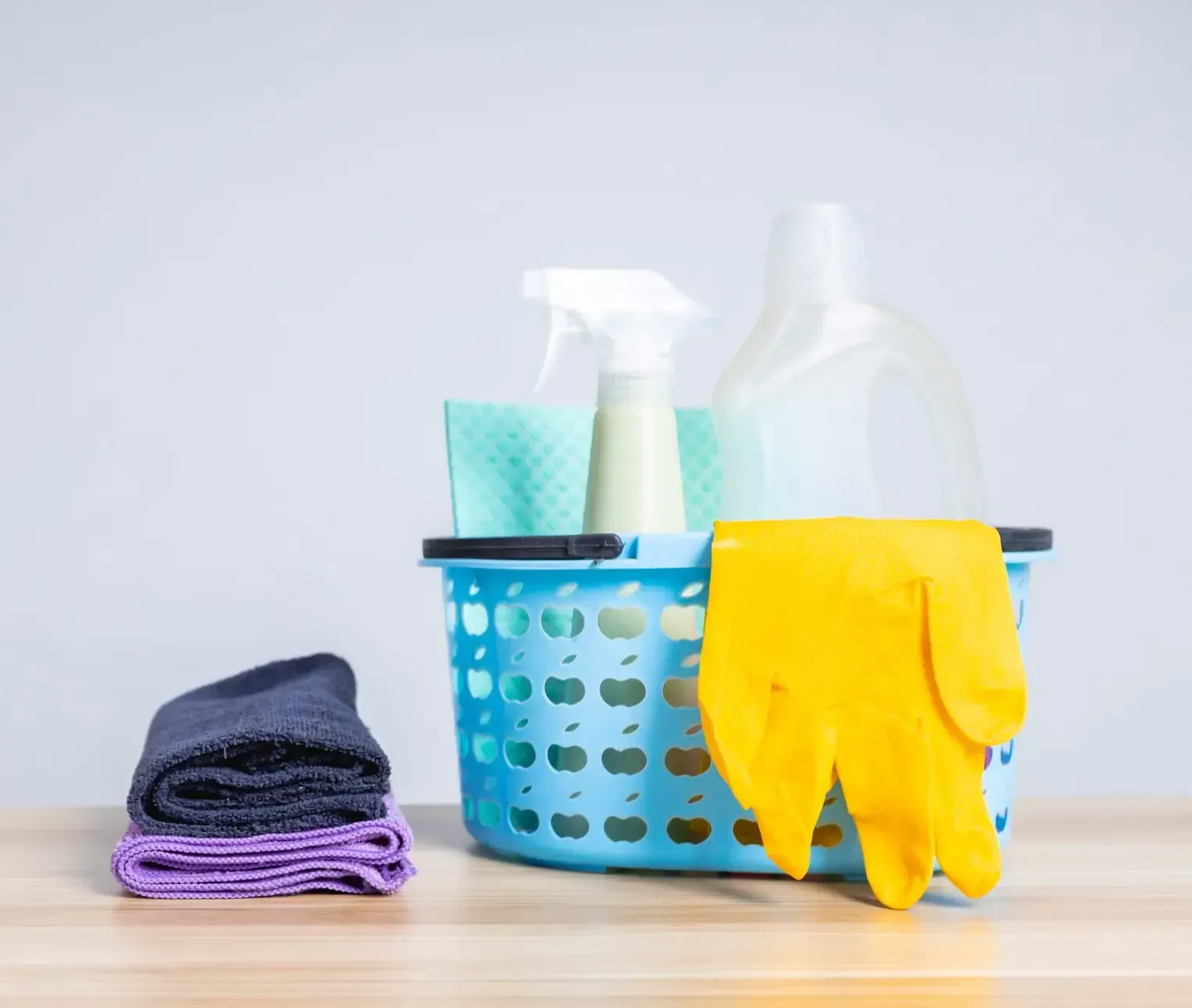 Una cesta de plástico azul que contenía productos de limpieza, guantes de goma amarillos y una botella pulverizadora, con dos paños doblados cerca.