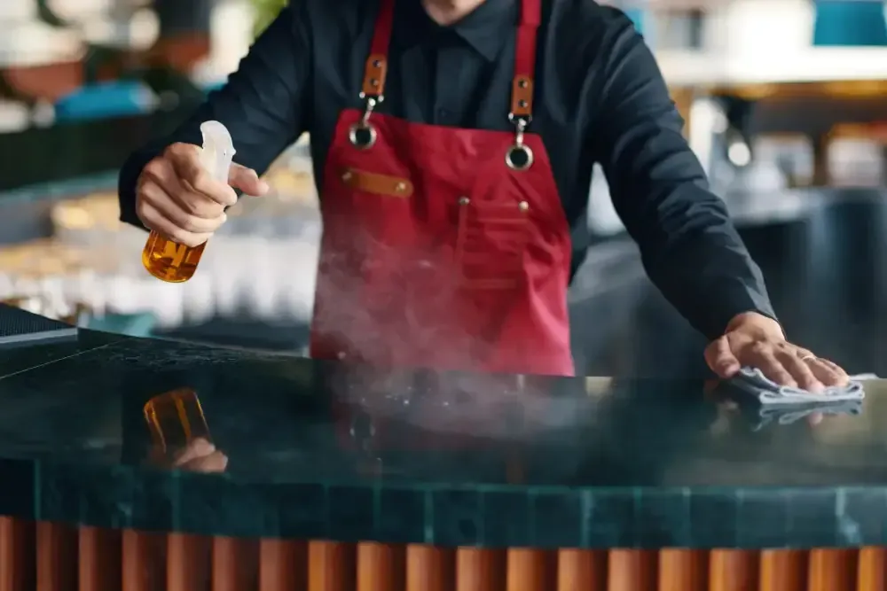 En un restaurante, una persona vestida con una camisa negra y un delantal rojo limpia una encimera oscura con un pulverizador y un paño.