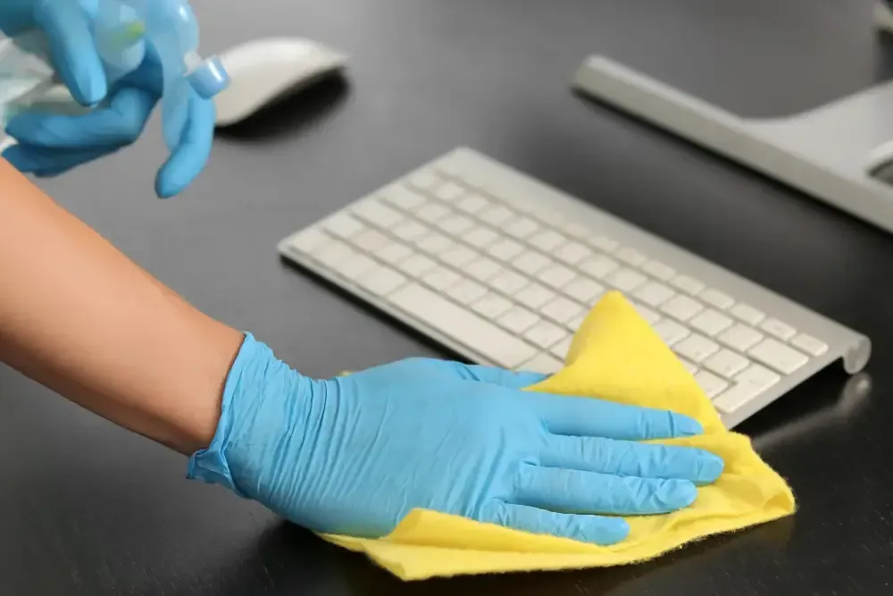 Una persona con guantes azules limpia un escritorio negro con un paño amarillo cerca de un teclado y un ratón de ordenador.