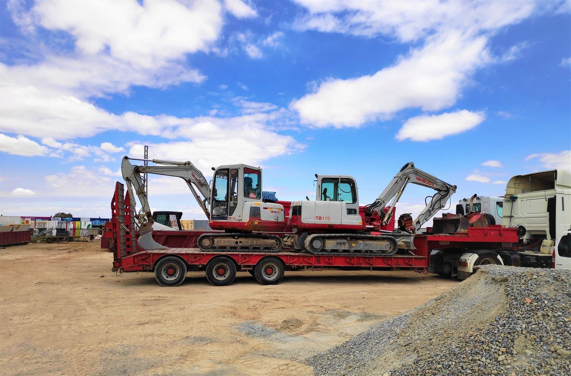 Dos excavadoras en un remolque rojo de plataforma, cargadas en un semirremolque. Con el cielo azul como fondo.
