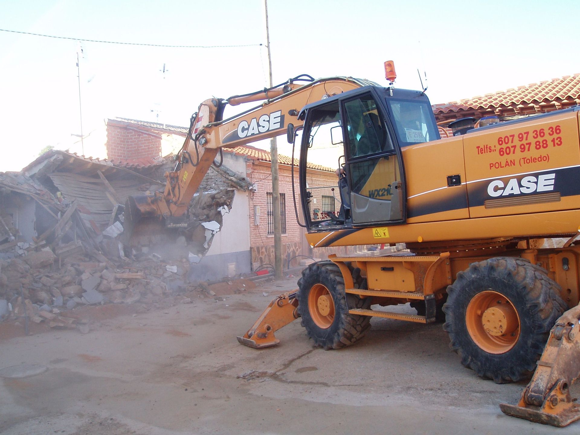 Excavadora CASE naranja demoliendo un edificio de ladrillos, calle polvorienta.