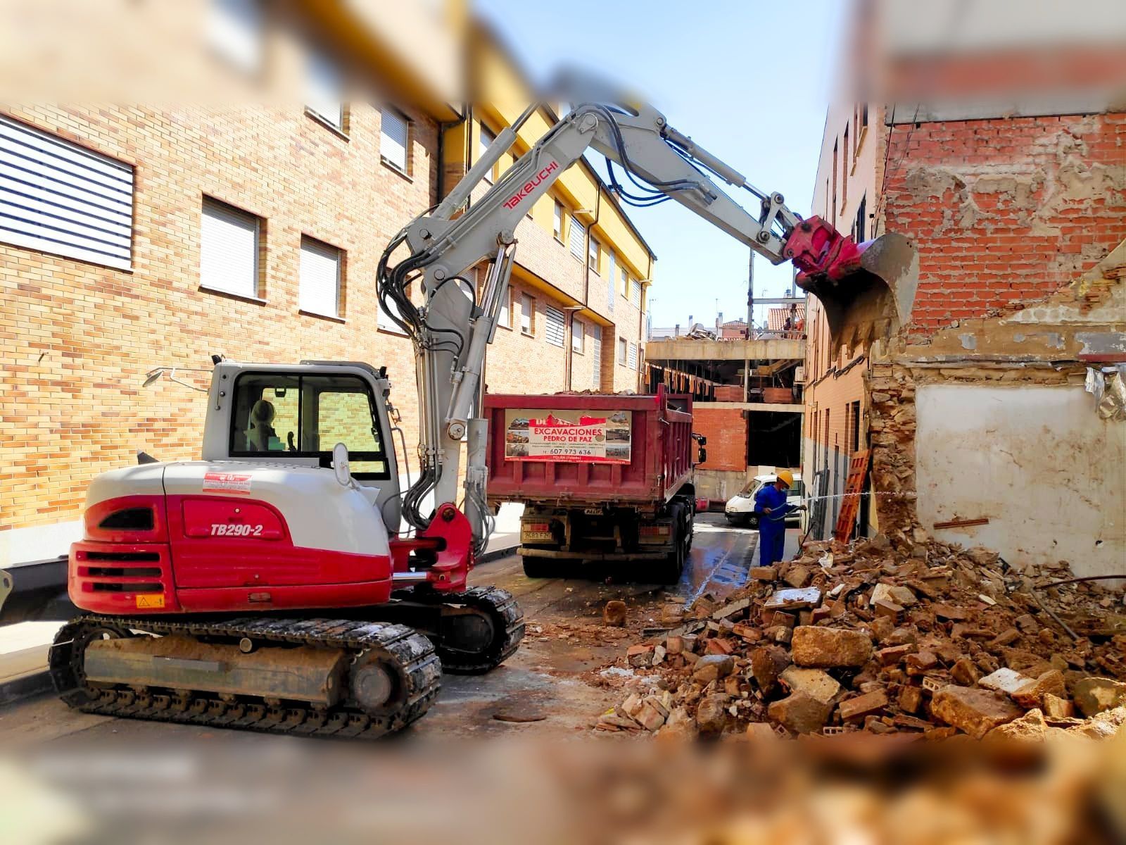 Una excavadora derriba un edificio y carga los escombros en un camión volquete. Obra al aire libre con edificios de ladrillo.