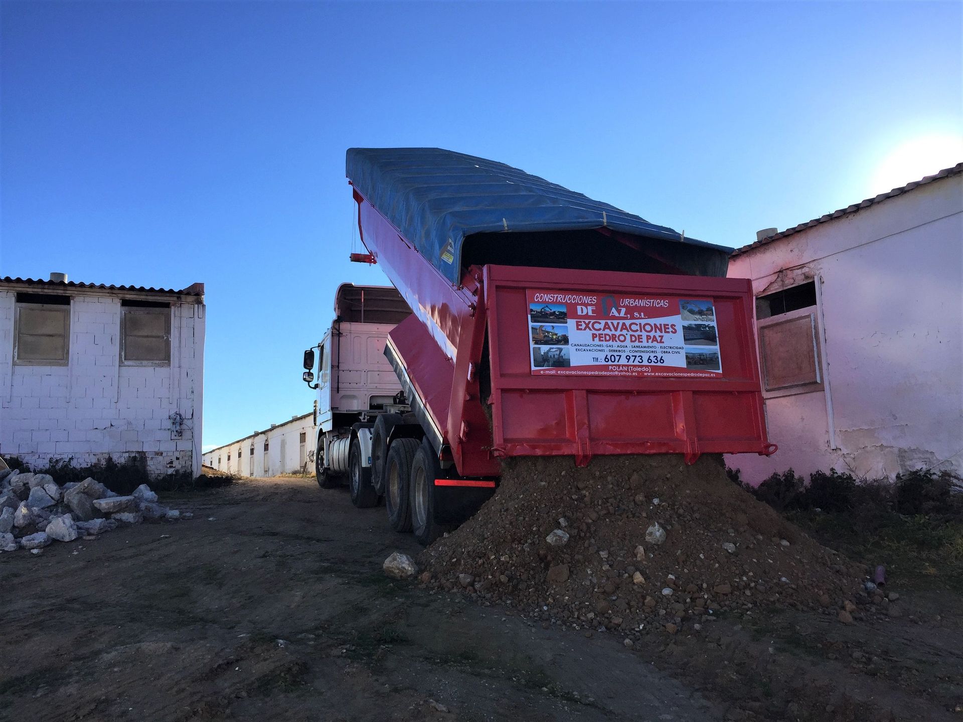 Camión volquete rojo descargando grava cerca de edificios blancos bajo un cielo azul.