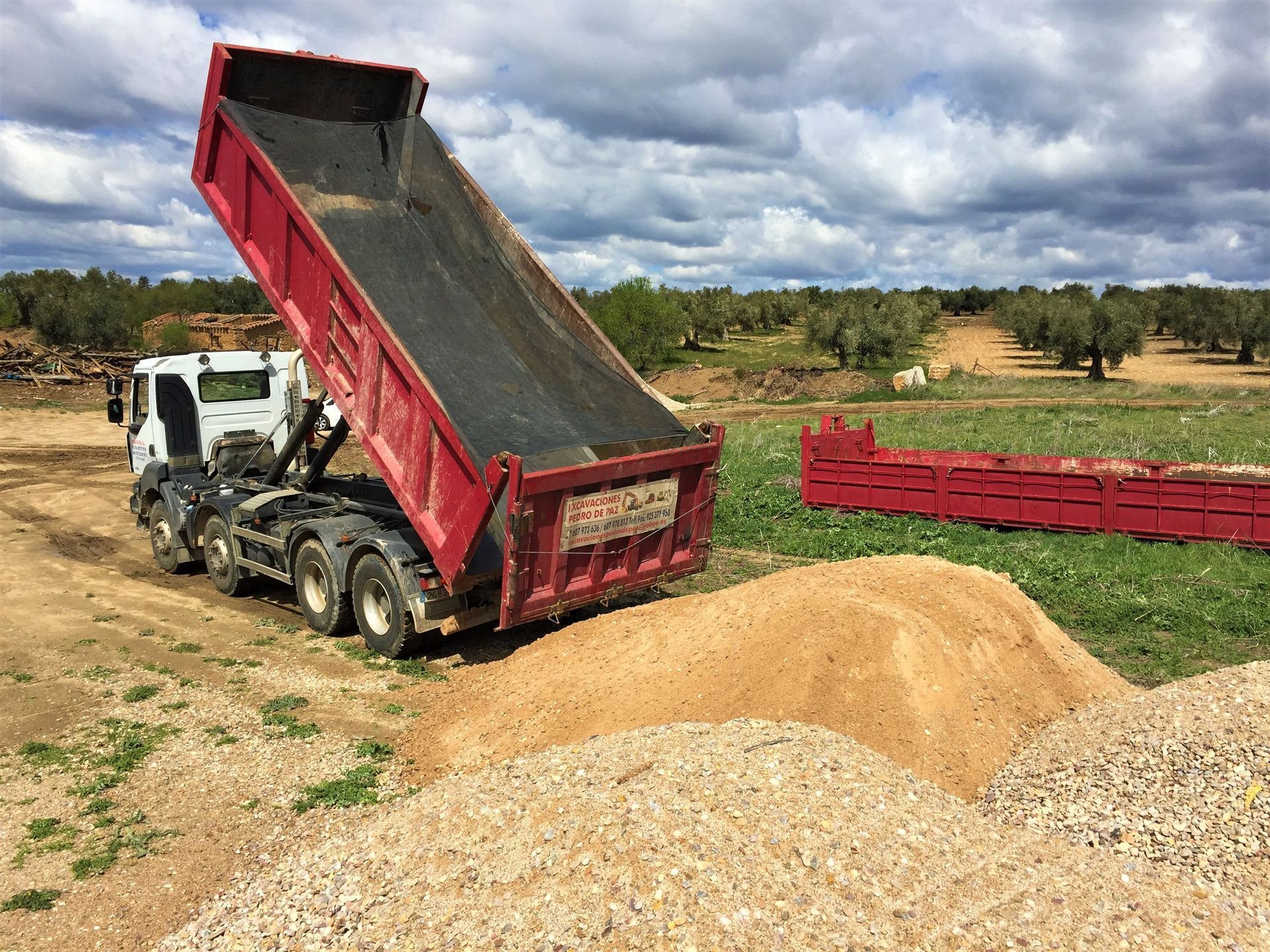 Camión volquete rojo descargando grava sobre una pila en un campo bajo un cielo nublado.