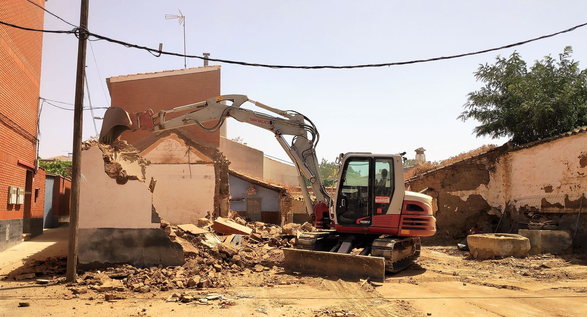 Excavadora demoliendo un edificio; escombros en el suelo; entorno soleado al aire libre.