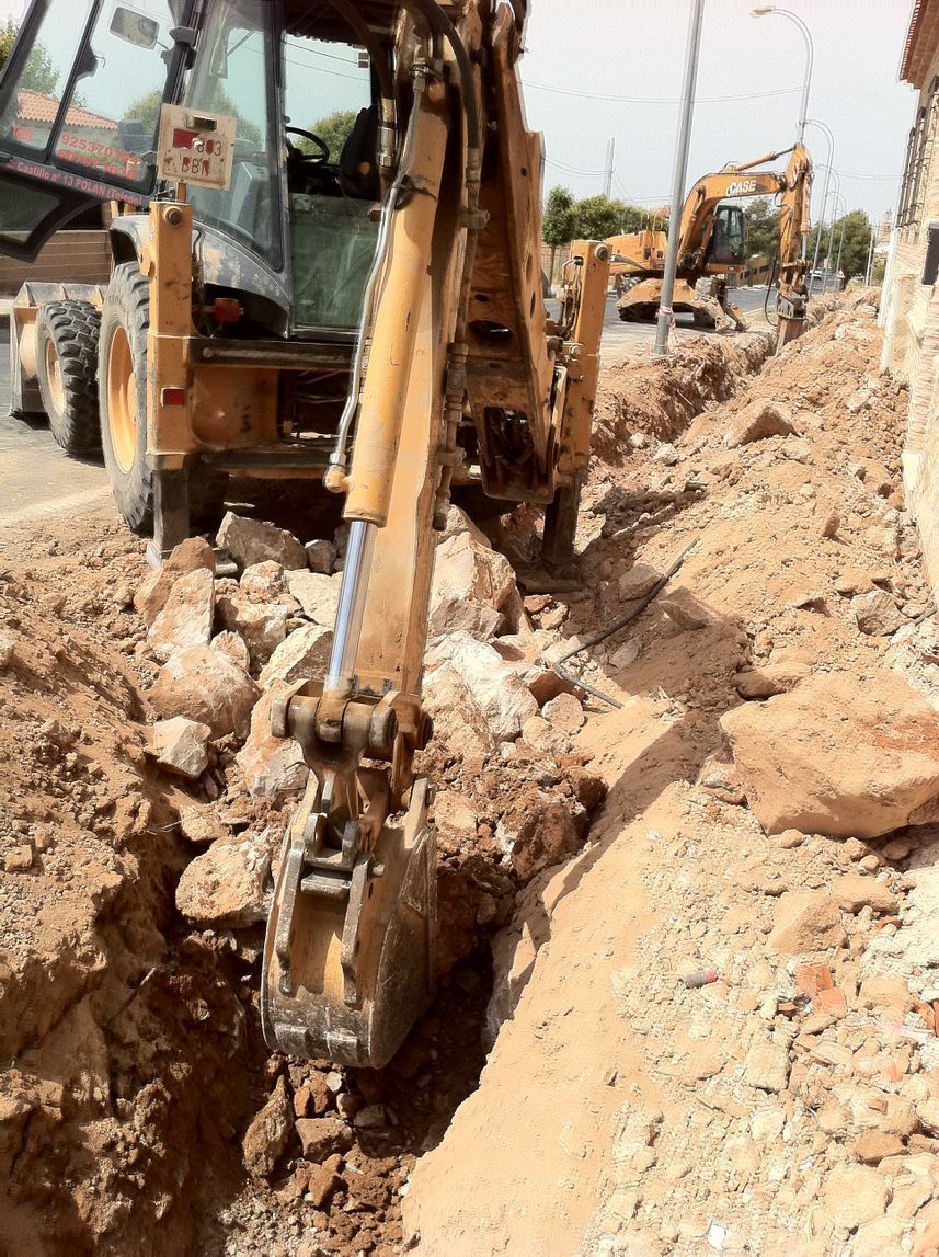 Retroexcavadora cavando una zanja al lado de una carretera, otros equipos de construcción en el fondo.