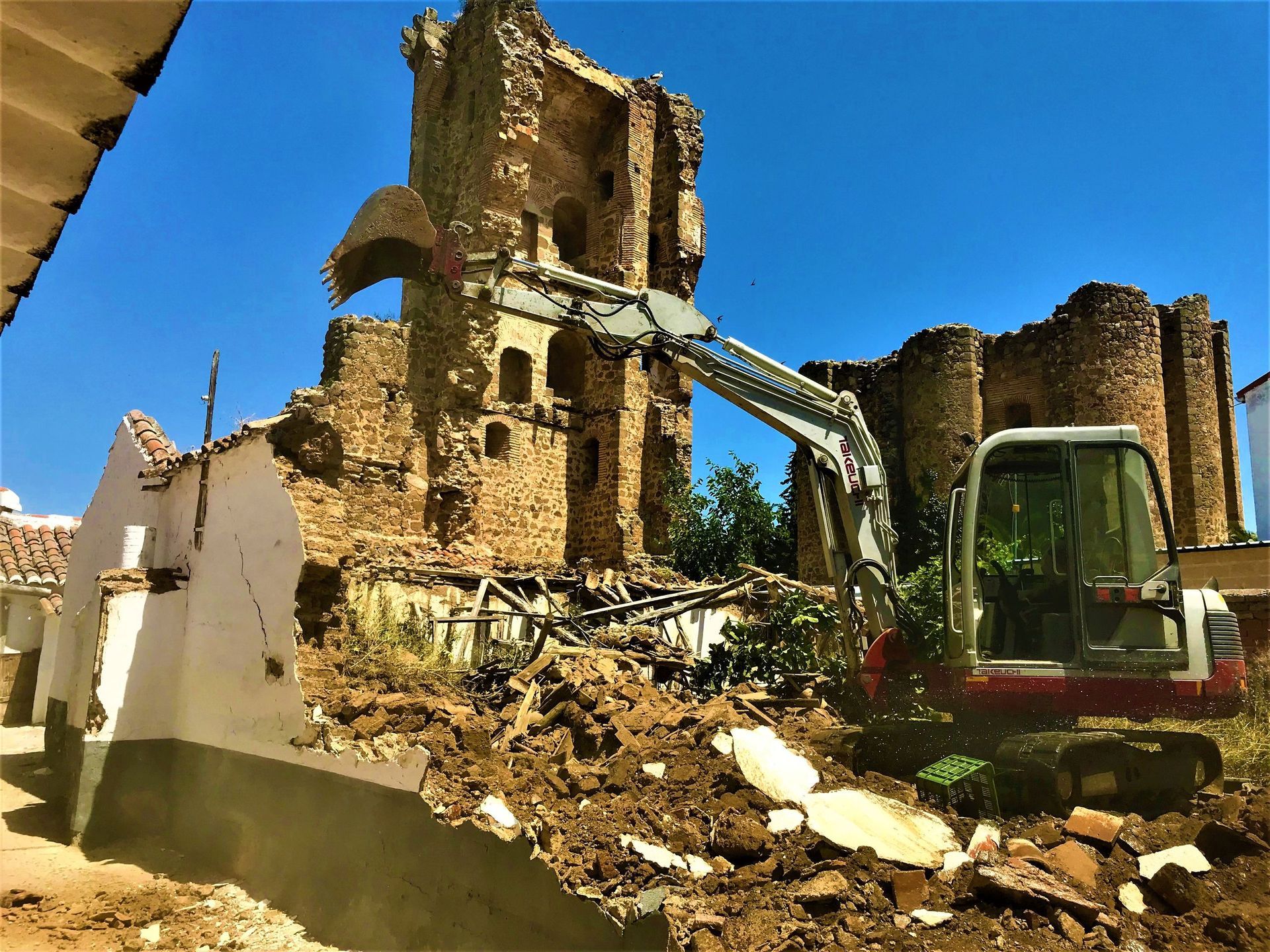 Excavadora demoliendo un edificio con una torre de piedra al fondo bajo un cielo azul.