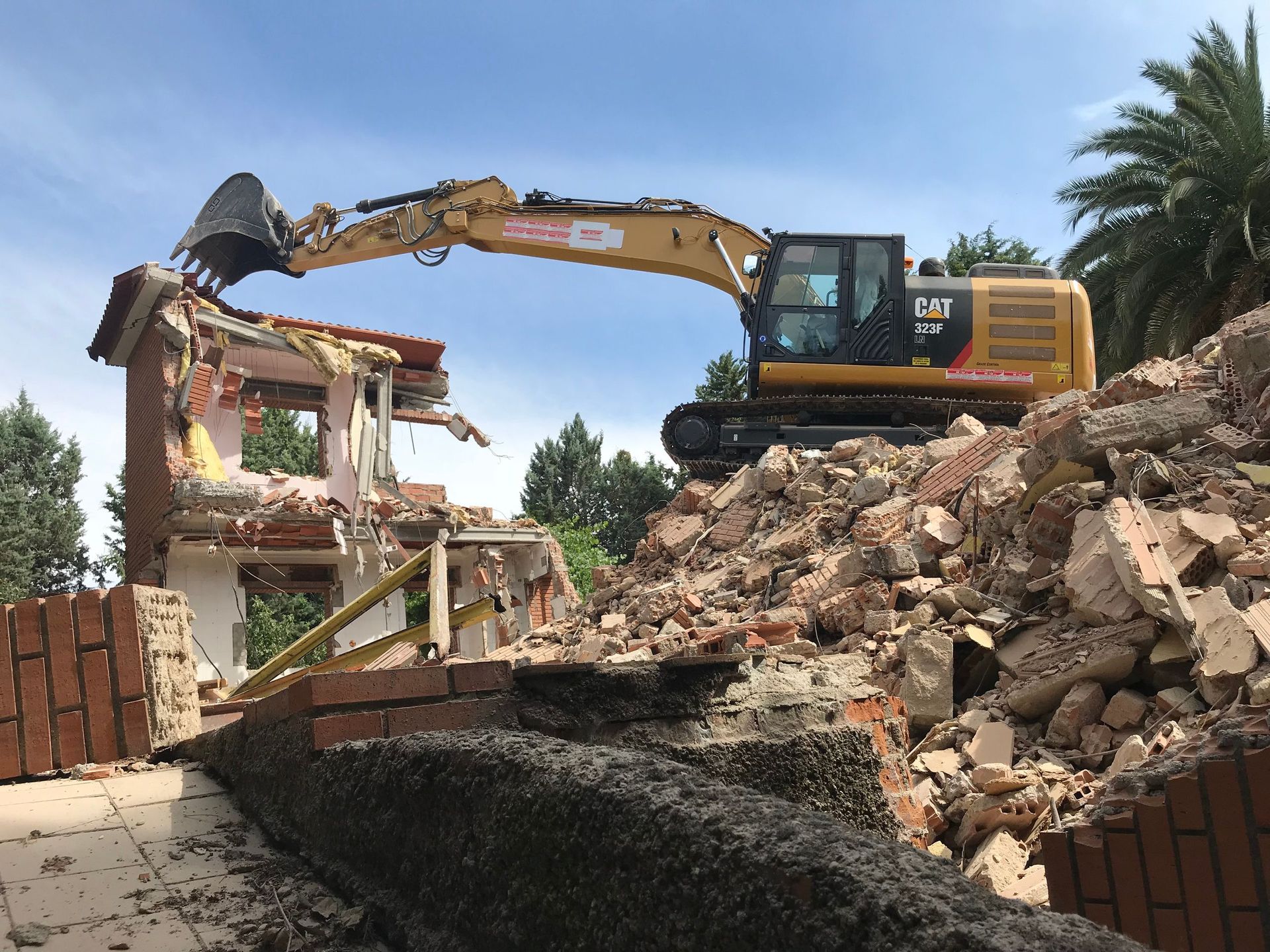 Excavadora demoliendo un edificio de ladrillos bajo un cielo azul, escombros en primer plano.