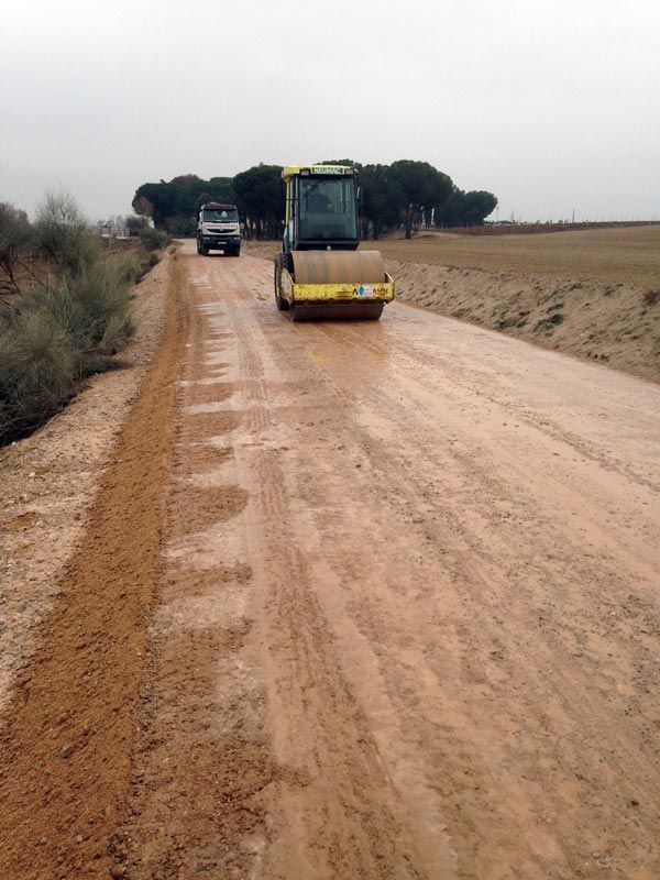 Construcción de carreteras: una apisonadora compacta el camino de grava; el camión la sigue, campo al fondo.