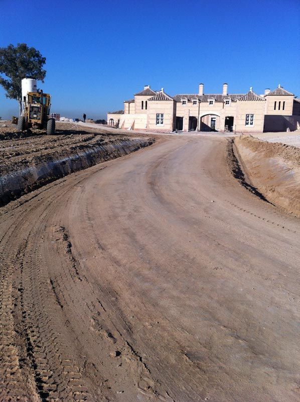 Camino de tierra que conduce a un edificio de color claro en construcción; maquinaria pesada a la izquierda.