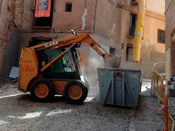 Cargadora compacta CASE de color naranja arrojando escombros a un contenedor de basura en un callejón adoquinado.