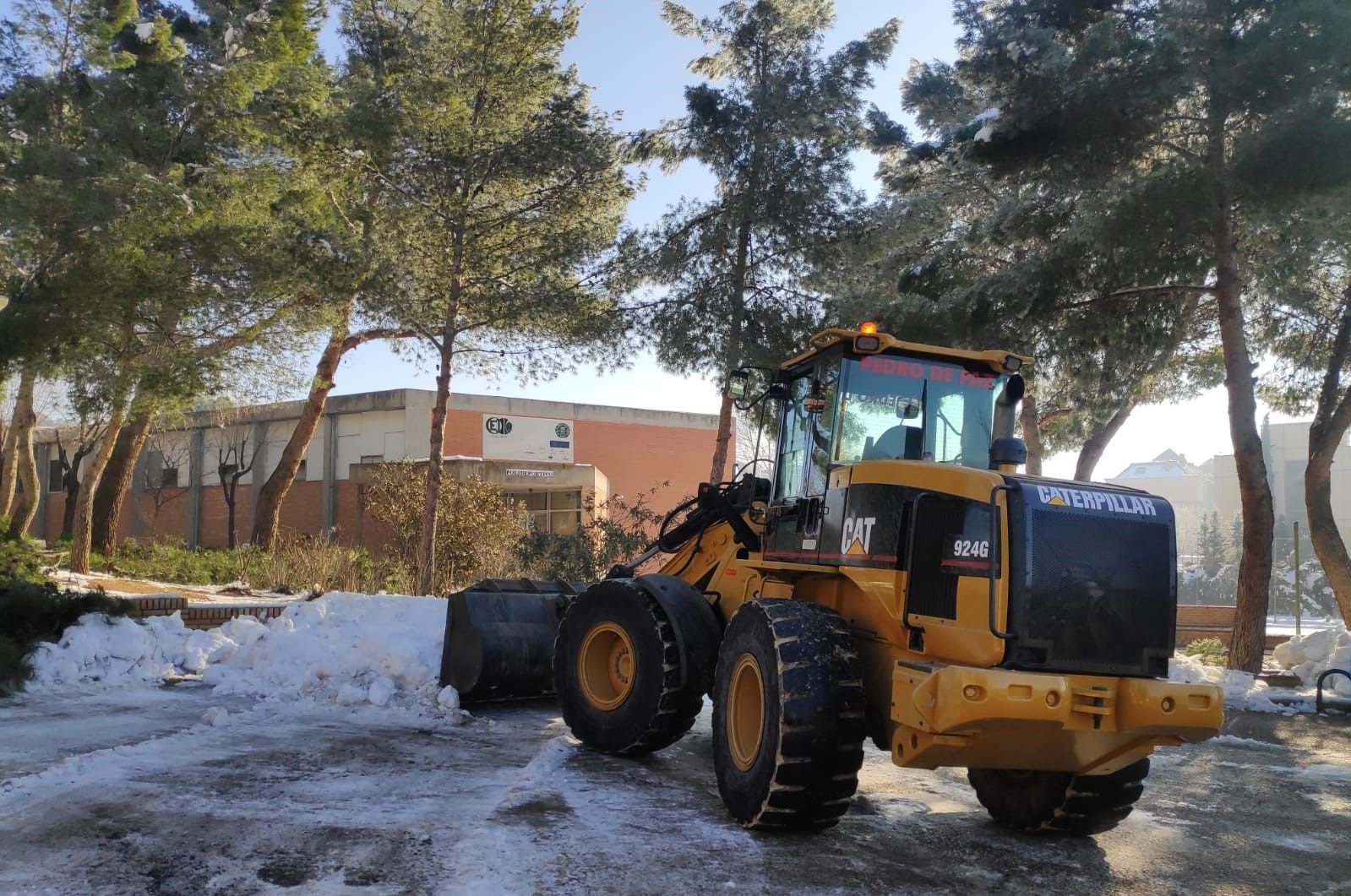 Cargadora de ruedas Caterpillar amarilla limpiando la nieve de una zona pavimentada cerca de árboles y un edificio en un día soleado.
