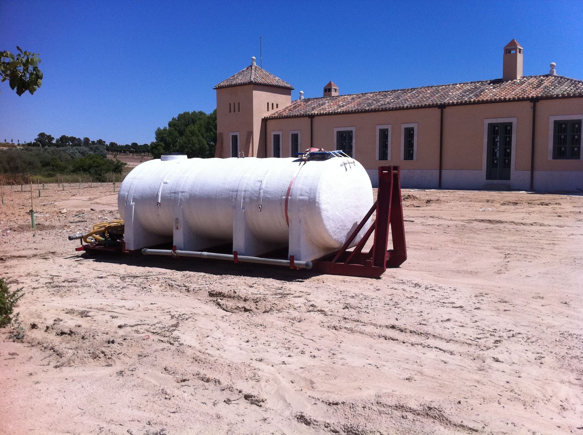 Tanque de agua blanca sobre marco de metal rojo en una zona arenosa, edificio al fondo.