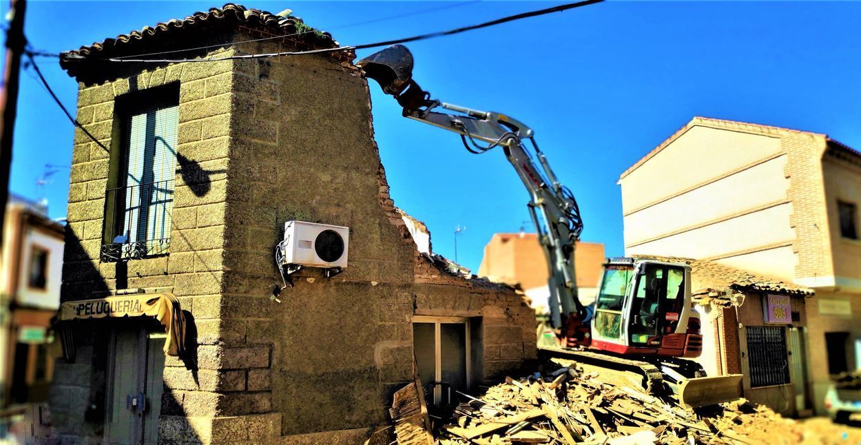 Excavadora demoliendo un edificio de piedra en un día soleado. Escombros y otros edificios visibles.