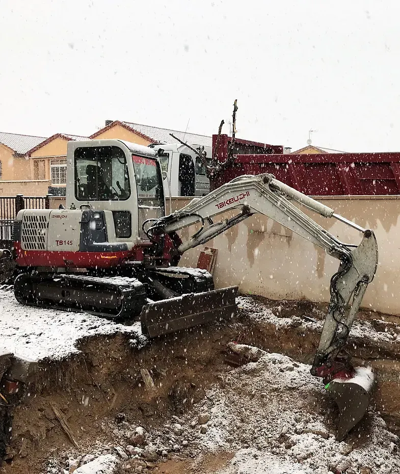 Miniexcavadora en la nieve, cavando cerca de una casa, con un camión al fondo.