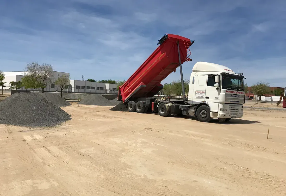 Camión volquete con caja roja descargando grava en un sitio de construcción.