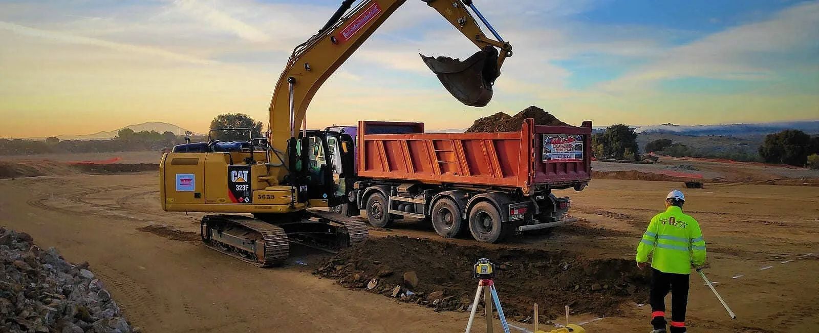 Una excavadora carga un camión volquete en un sitio de construcción; un trabajador con ropa de alta visibilidad camina en primer plano.