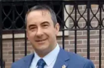 Un hombre con traje y corbata sonríe delante de una valla.