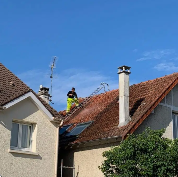 Une personne nettoie à haute pression un toit de tuiles rouges ; ciel bleu dégagé au-dessus d'une maison avec une cheminée et une antenne.