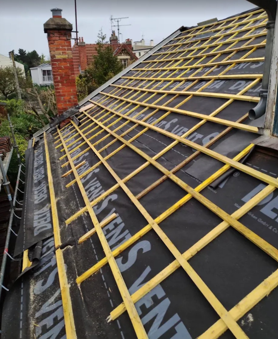 Toiture partiellement recouverte d'une sous-couche noire et d'une charpente en bois jaune, cheminée en arrière-plan.