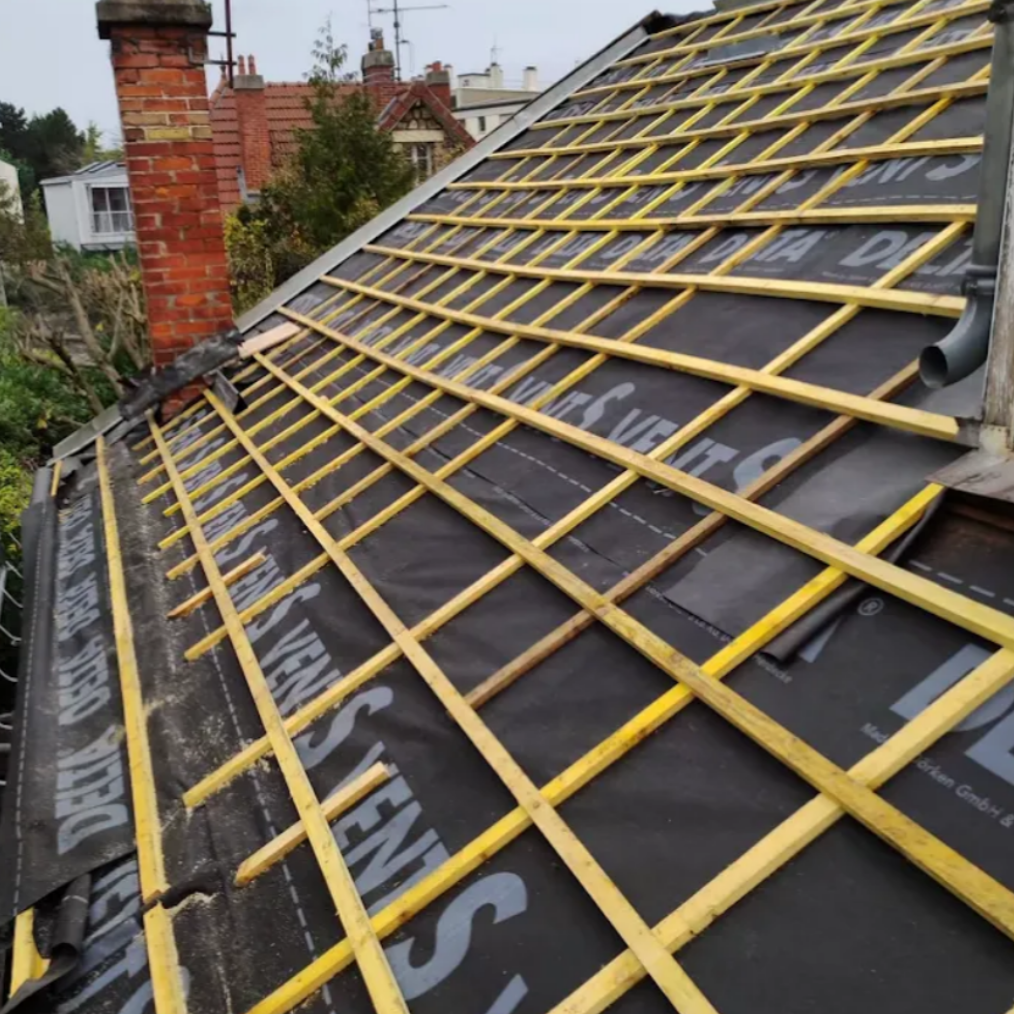 Toiture en construction : charpente en bois sur sous-toiture noire. Cheminée en briques, ciel couvert.