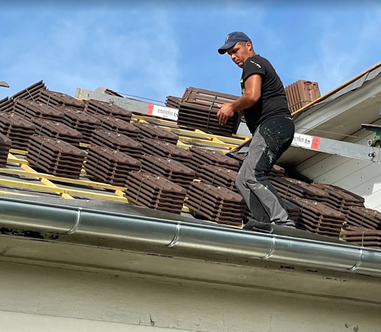 Un couvreur transporte des tuiles sur un toit. Il porte une casquette et des vêtements sombres. Des tuiles brunes sont empilées sur le toit.
