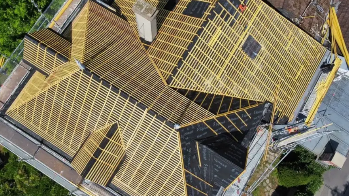 Vue aérienne d'un toit en construction ; des poutres en bois et des matériaux de couverture noirs recouvrent la structure.