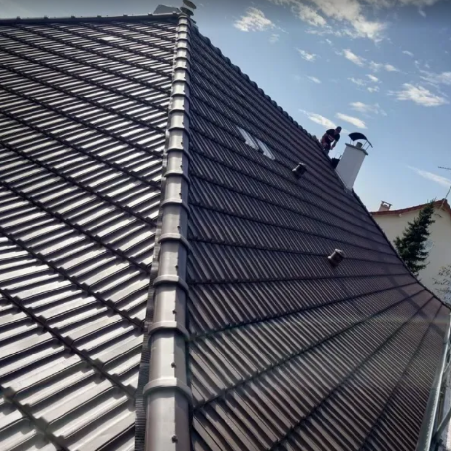 Toit de tuiles brun foncé avec des ouvriers, cheminée blanche, ciel bleu.