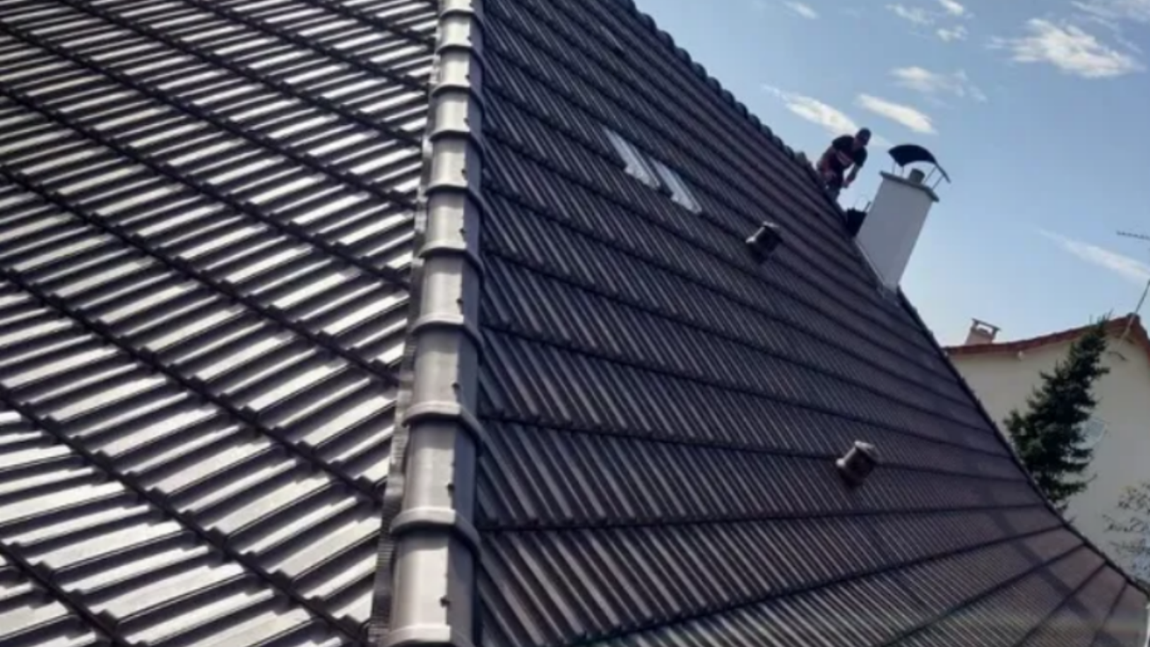 Toit de tuiles brun foncé avec deux personnages au travail, cheminée, ciel bleu.