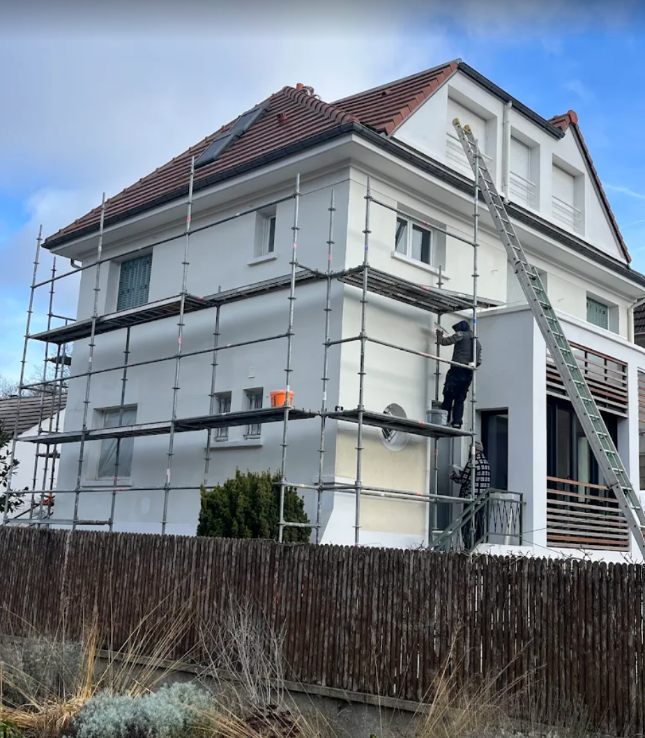 Des échafaudages entourent une maison blanche à deux étages sur laquelle se trouve une personne. Une échelle est appuyée contre le mur.