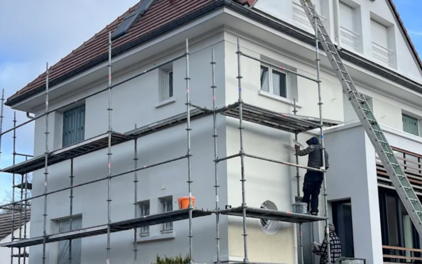 Échafaudages autour d'une maison à deux étages, une personne en train de peindre l'extérieur, ciel couvert.