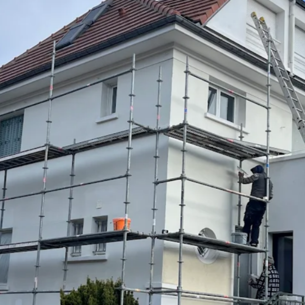 Un homme sur un échafaudage peint la façade d'un bâtiment blanc. Un seau orange est visible.
