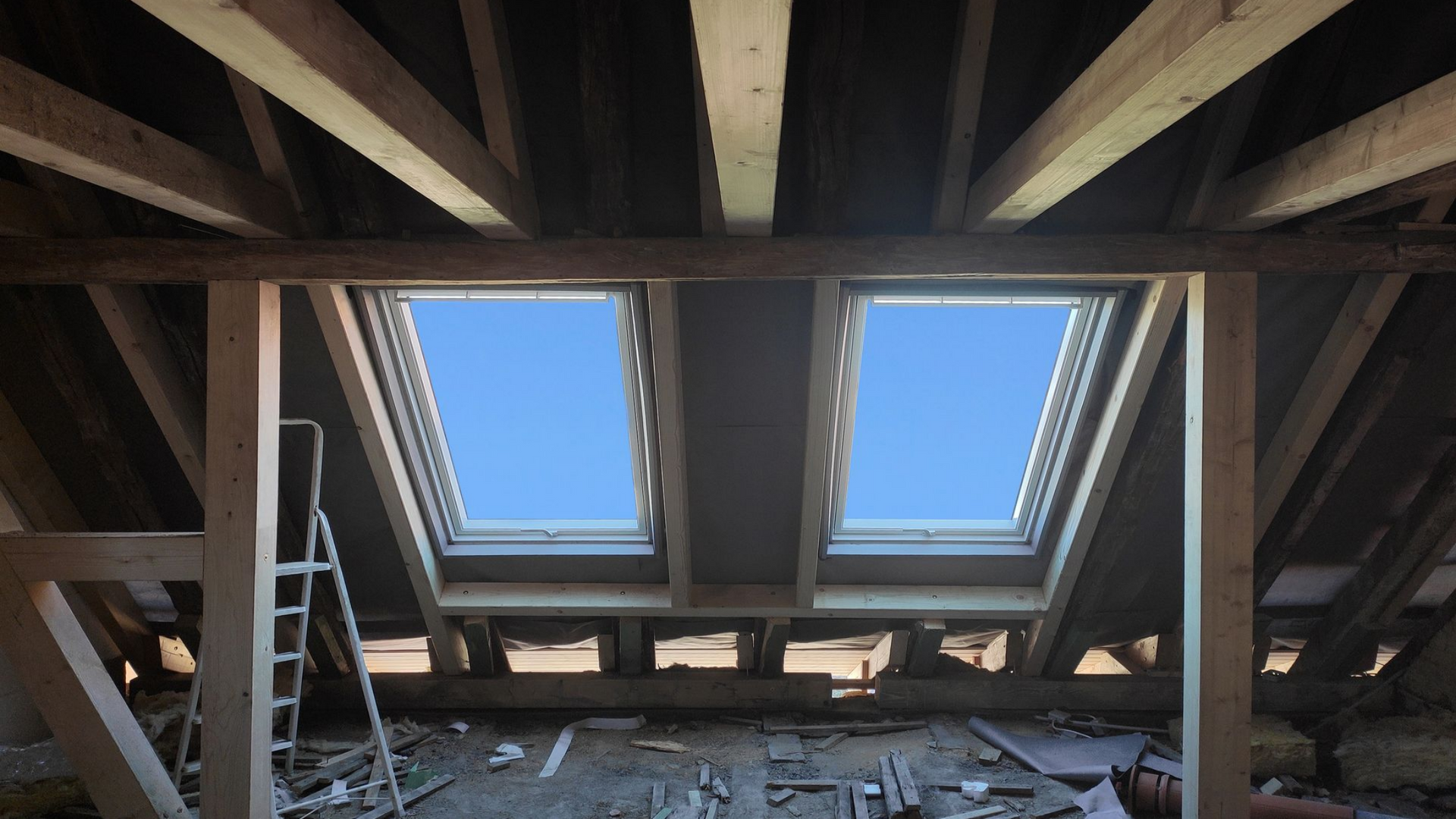 Vue intérieure d'un grenier avec deux puits de lumière laissant apparaître un ciel bleu éclatant, des poutres en bois et une échelle.