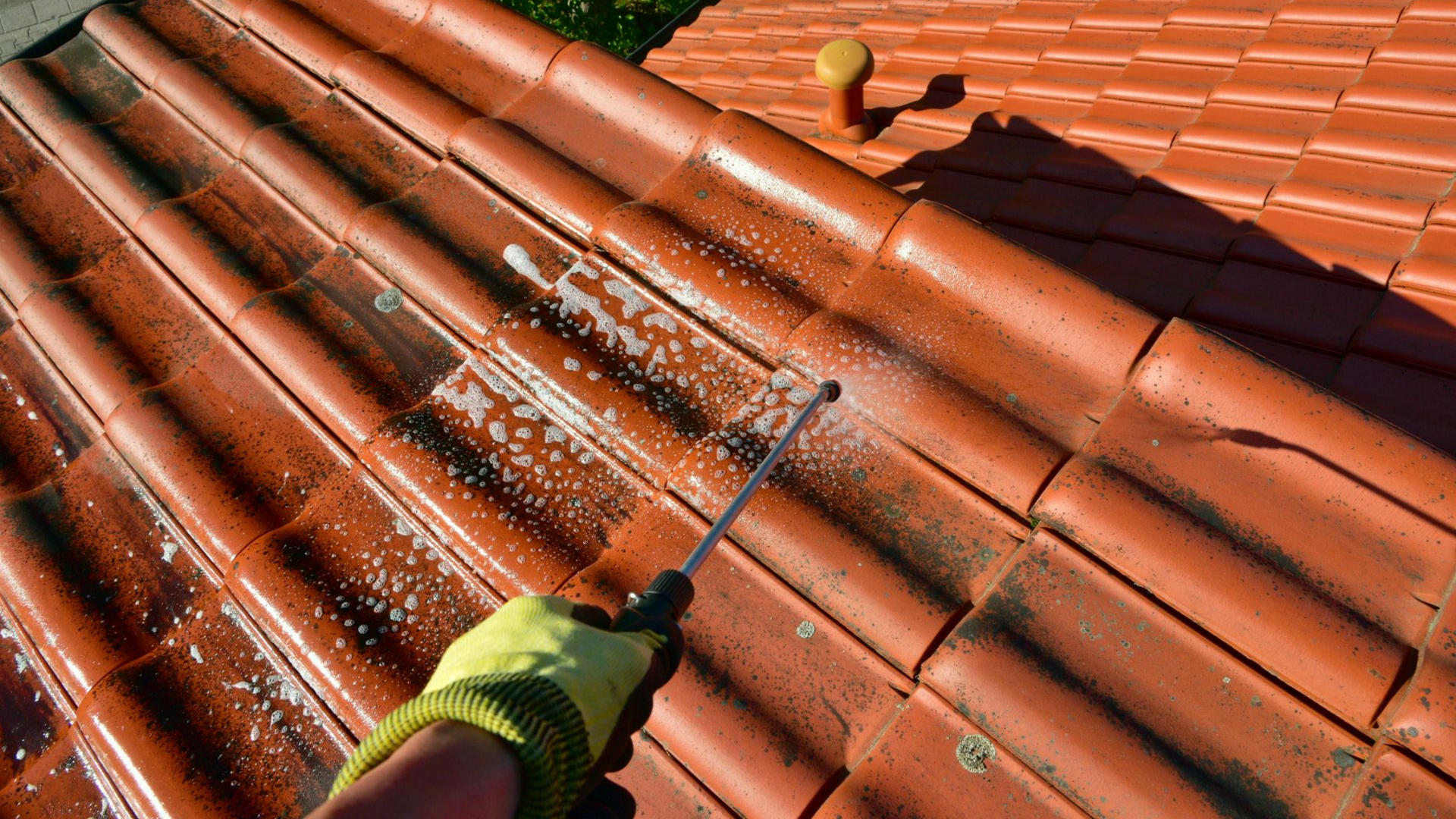 Une personne utilise un nettoyeur à haute pression pour nettoyer un toit de tuiles rouges, portant un gant jaune.