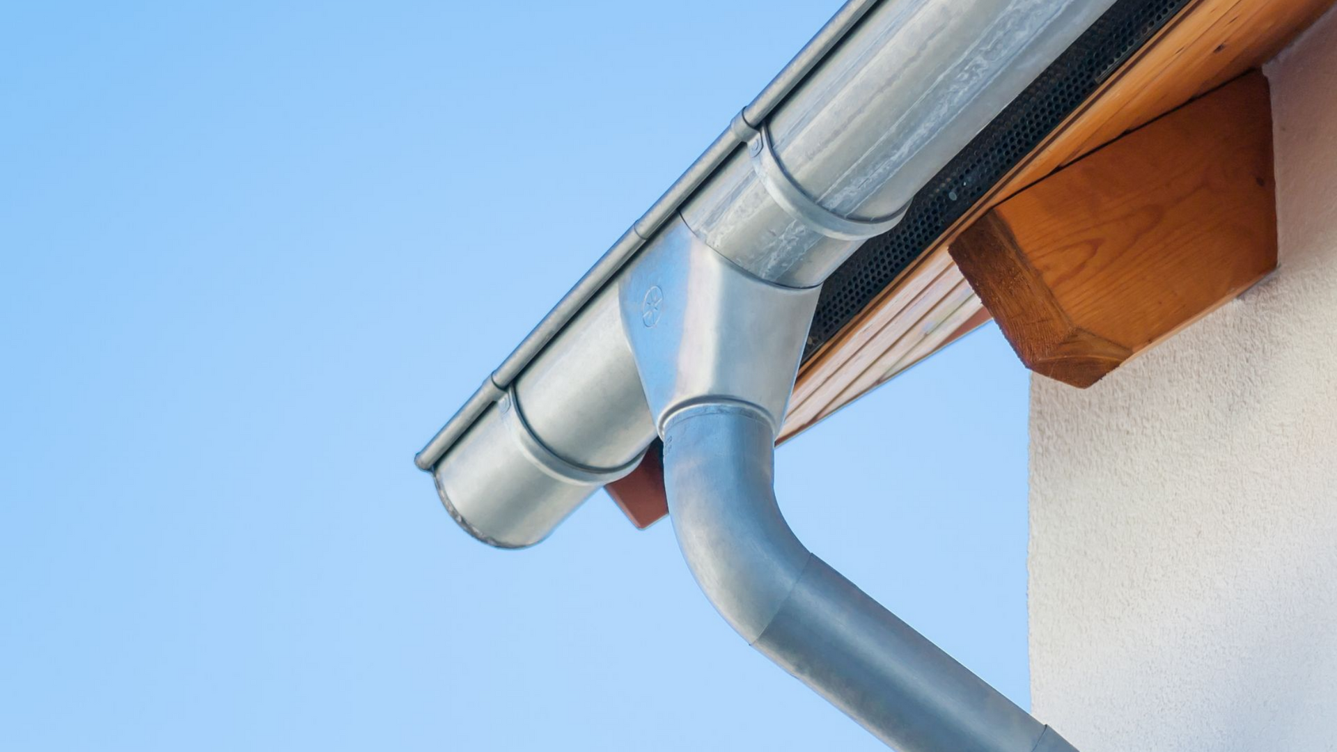 Gros plan sur une gouttière en métal argenté, avec une descente pluviale sur fond de ciel bleu.