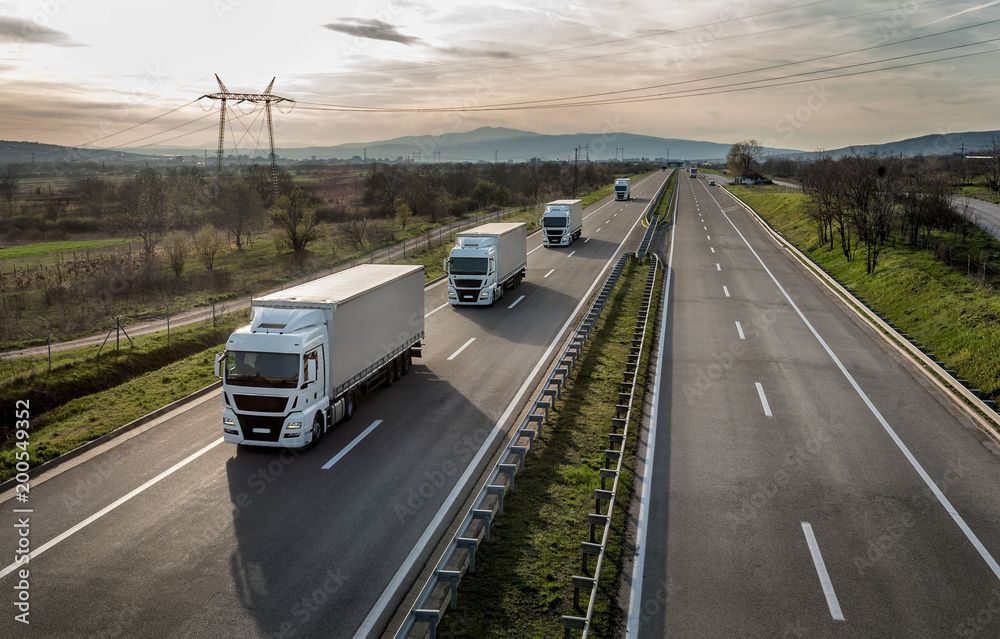 Drei Lastwagen fahren auf einer mehrspurigen Autobahn unter bewölktem Himmel, Felder und Stromleitungen in der Nähe.
