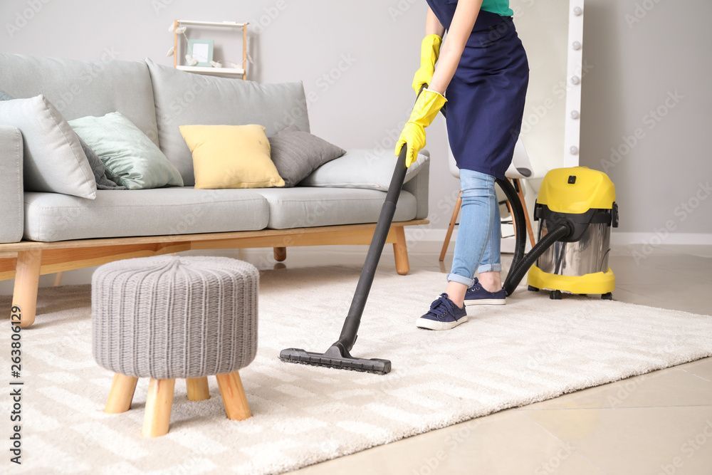 Eine Person saugt einen weissen Teppich in einem hellen Wohnzimmer mit grauem Sofa und gelbem Staubsauger.