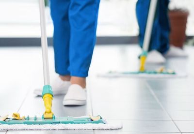 Eine Person, die mit einem Wischmopp einen Fliesenboden wischt und dabei eine blaue Hose und weisse Schuhe trägt.