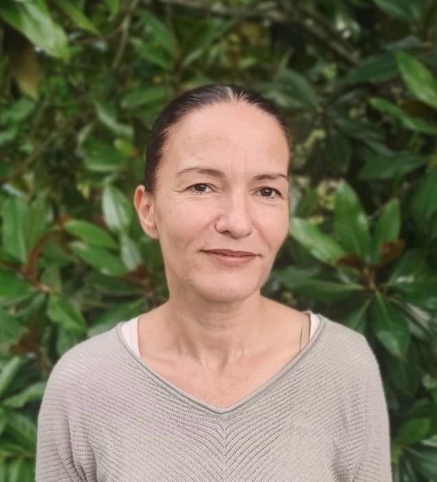 Mujer con cabello castaño con un suéter ligero, mirando a la cámara; fondo de follaje verde.