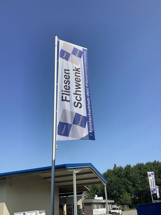 Flagge „Fliesen Schwenk“, weiß mit blauer Schrift und Logo, weht vor dem Gebäude.