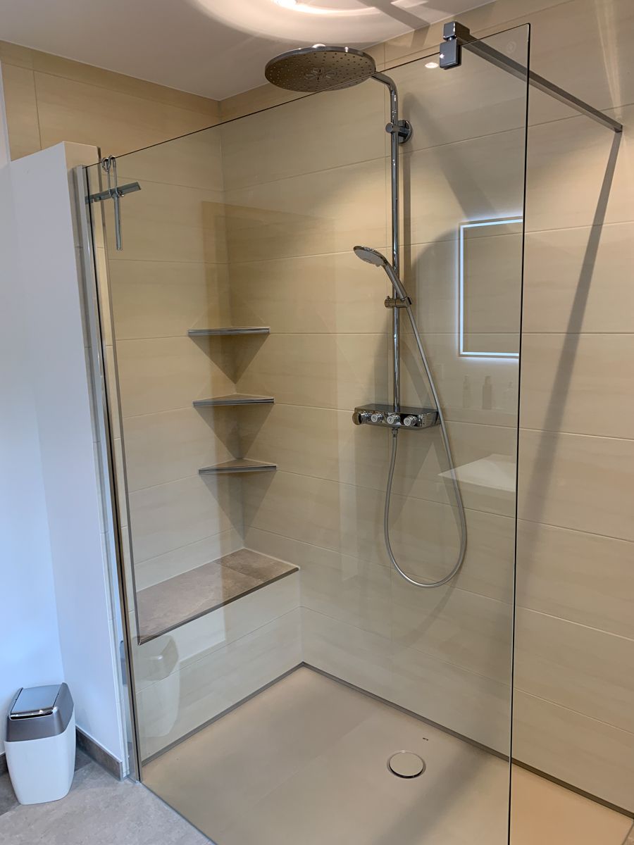 Dusche mit Glasabtrennung, integriertem Sitz, drei Eckablagen und Regenduschkopf. Beige Fliesenwände.